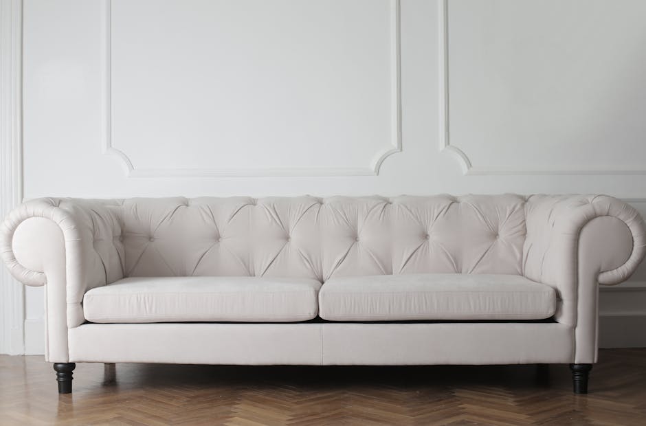 A person using a handheld vacuum cleaner to deep clean a decorative cushion with a yellow and white geometric pattern, placed on a dark fabric sofa in a well-lit room, demonstrating surface cleaning a