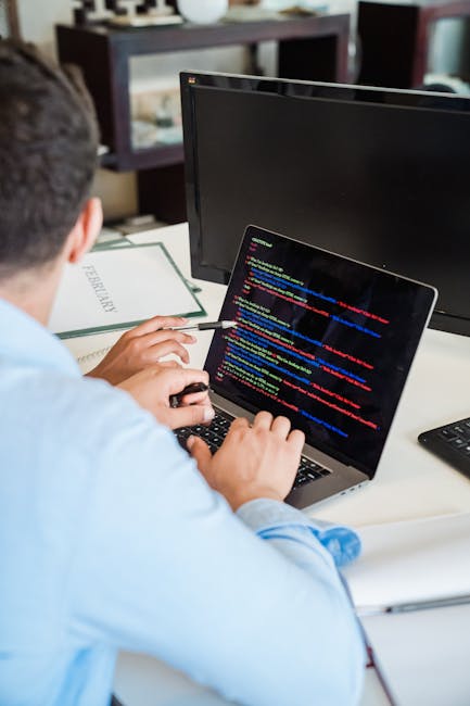 A person sitting at a desk working on a laptop with a black screen displaying colorful lines of code. The desk contains a closed book titled 'PERFECTION,' a white sheet of paper, and a desk mouse. In 