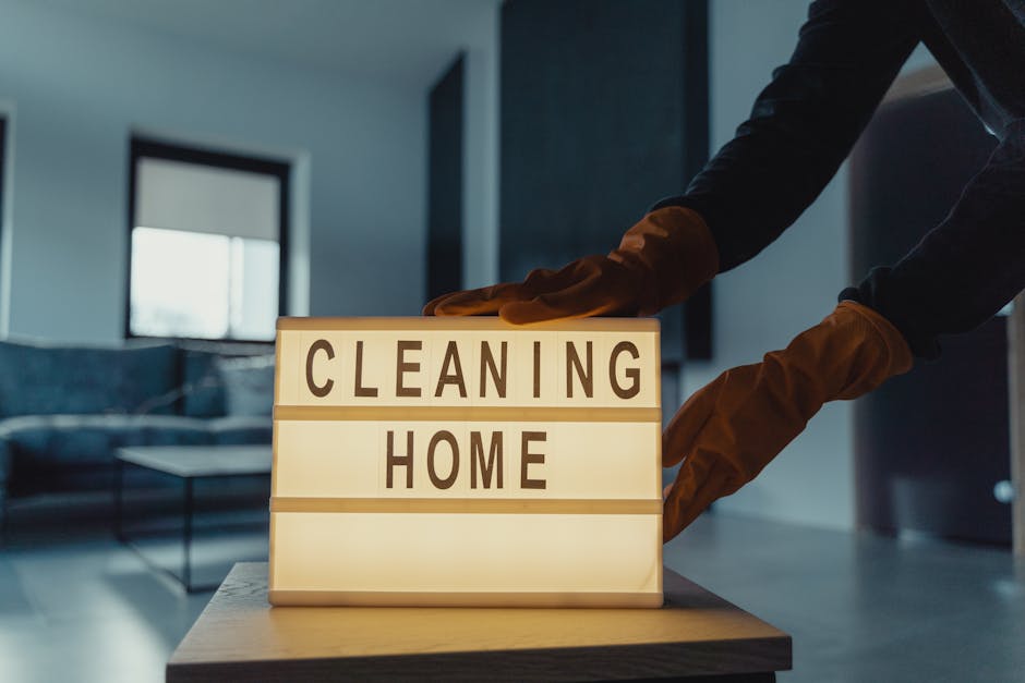 A person wearing orange gloves arranging a white lightbox with the words 'CLEANING HOME' in bold black letters on a wooden table in a modern, well-lit living room with large windows and contemporary f